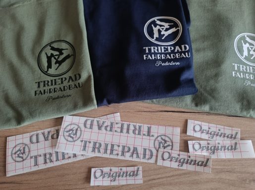T-Shirt Triepad Fahrradbau Paderborn Drei Textilien in verschiedenen Farben und Aufkleber mit "TRIPAD" und "Original".
